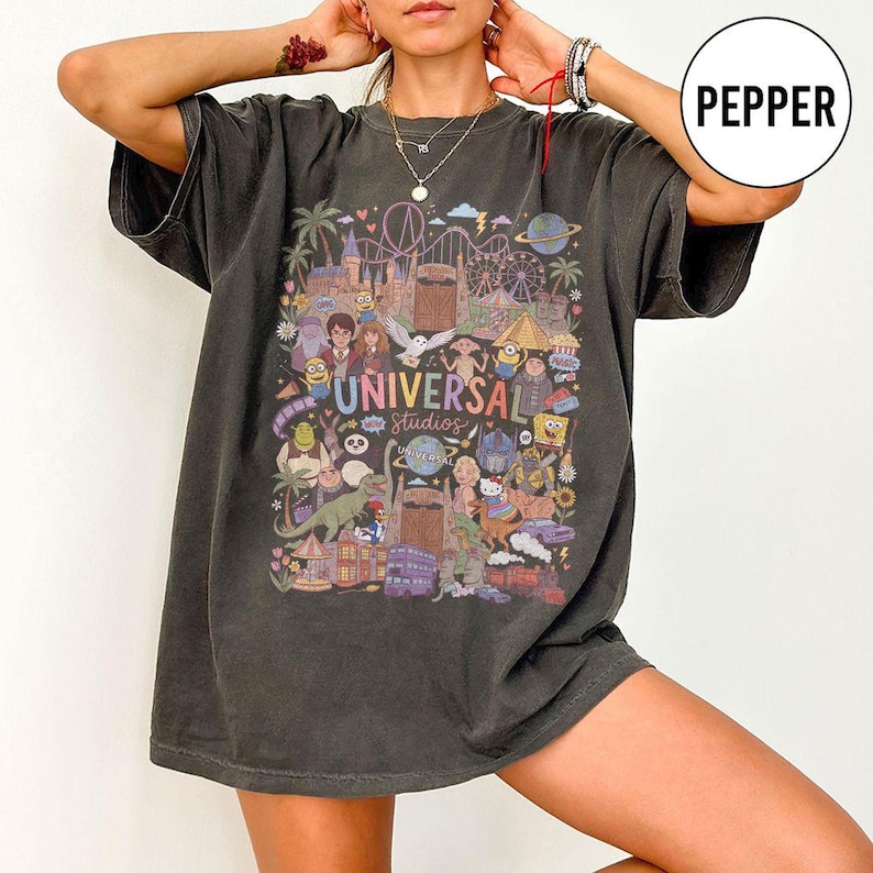 Może przedstawiać: Ciemnoszary t-shirt o kroju oversize z kolorowym nadrukiem graficznym przedstawiającym atrakcje i postacie z Universal Studios, w tym Harry'ego Pottera, Shreka i Minionki, z napisem Universal Studios na środku wzoru.
