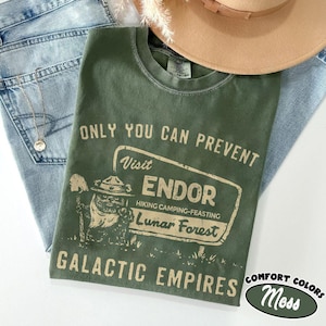 Puede incluir: Una camiseta verde musgo con el texto "ONLY YOU CAN PREVENT Visit ENDOR Hiking Camping-Feasting Lunar Forest GALACTIC EMPIRES". La camiseta se combina con vaqueros azul claro y un sombrero marrón.