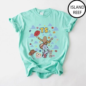 May include: A mint green t-shirt featuring a graphic print of a person with long hair wearing a yellow plaid shirt and cow-print pants, holding a guitar. The design includes a red hat, a brown cowboy hat, musical notes, clouds, and the text TS.