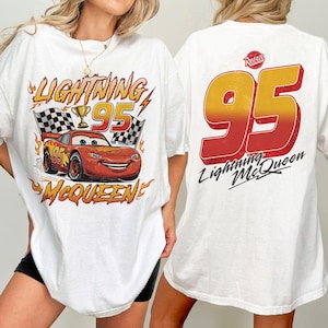 K&ouml;nnte beinhalten: Wei&szlig;es T-Shirt mit einer Grafik von Lightning McQueen aus dem Film Cars. Das Design auf der Vorderseite zeigt den Text "Lightning 95 McQueen" mit einem Cartoon-Auto, karierten Flaggen und einer Troph&auml;e. Auf der R&uuml;ckseite steht gro&szlig; "95" und "Lightning McQueen".