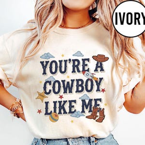 Puede incluir: Una camiseta de color marfil con el texto azul oscuro "YOU'RE A COWBOY LIKE ME" rodeado de ilustraciones gr&aacute;ficas de un sombrero de vaquero marr&oacute;n, un cohete, una estrella de sheriff, una pelota, peque&ntilde;as estrellas rojas, estrellas amarillas y nubes azul claro.