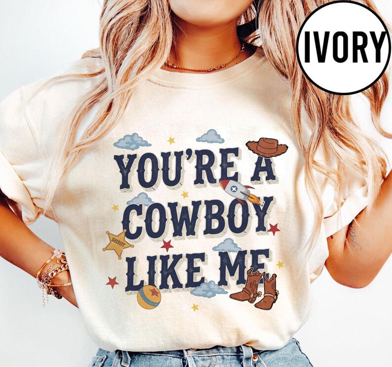 Puede incluir: Una camiseta de color marfil con el texto azul oscuro "YOU'RE A COWBOY LIKE ME" rodeado de ilustraciones gr&aacute;ficas de un sombrero de vaquero marr&oacute;n, un cohete, una estrella de sheriff, una pelota, peque&ntilde;as estrellas rojas, estrellas amarillas y nubes azul claro.