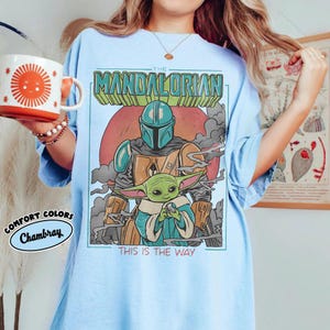 May include: Light blue t-shirt featuring a graphic of The Mandalorian and Grogu, with the text "THE MANDALORIAN" and "THIS IS THE WAY." The shirt is a Comfort Colors Chambray. A red and orange mug with a sun design is held in the image.