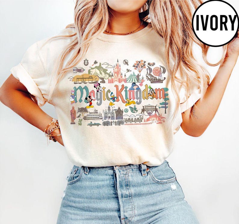 Op de afbeelding: Ivoor T-shirt met een kleurrijke Magic Kingdom-afbeelding. Het ontwerp bevat stripfiguren en parkherkenningspunten. Het shirt is gemaakt van zacht materiaal en heeft korte mouwen.