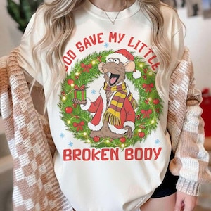 May include: Cream-colored graphic t-shirt featuring a cartoon rat in a Santa hat and scarf, holding a gift box within a Christmas wreath. The text reads "GOD SAVE MY LITTLE BROKEN BODY" in red.