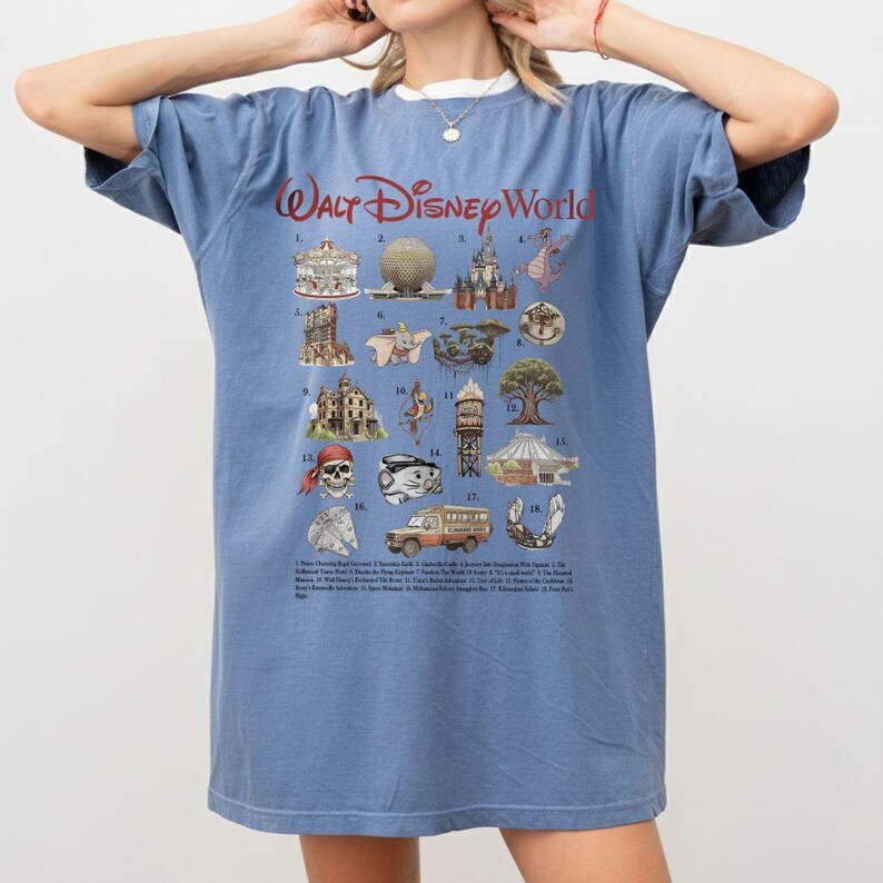 Puede incluir: Camiseta azul con el texto "Walt Disney World" en rojo. La camiseta presenta una cuadr&iacute;cula de ilustraciones coloridas de iconos de los parques Disney, incluyendo un carrusel, Spaceship Earth y un drag&oacute;n. Una lista de atracciones est&aacute; impresa debajo.