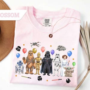May include: A light pink Comfort Colors crew neck sweatshirt featuring Star Wars characters holding balloons. The graphic includes R2-D2, Chewbacca, C-3PO, Darth Vader, a Stormtrooper, Princess Leia, and Baby Yoda. The word "Blossom" is in the upper left corner.