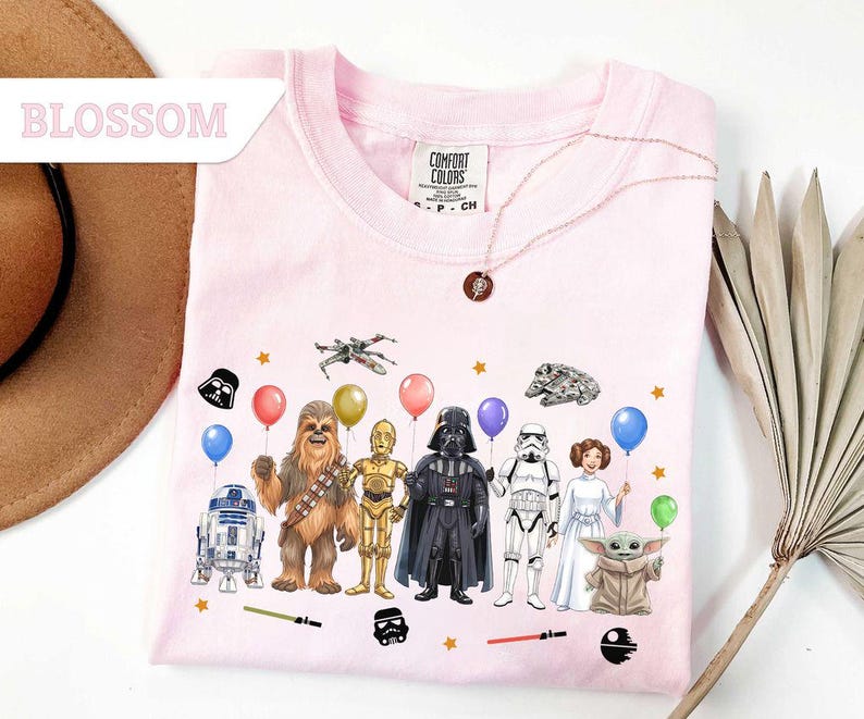 May include: A light pink Comfort Colors crew neck sweatshirt featuring Star Wars characters holding balloons. The graphic includes R2-D2, Chewbacca, C-3PO, Darth Vader, a Stormtrooper, Princess Leia, and Baby Yoda. The word "Blossom" is in the upper left corner.