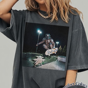 May include: A charcoal gray t-shirt featuring a graphic print of a character in metallic armor skateboarding in the air above a small green creature on a skateboard in a concrete park at night, with the text Comfort Colors Pepper in the corner.