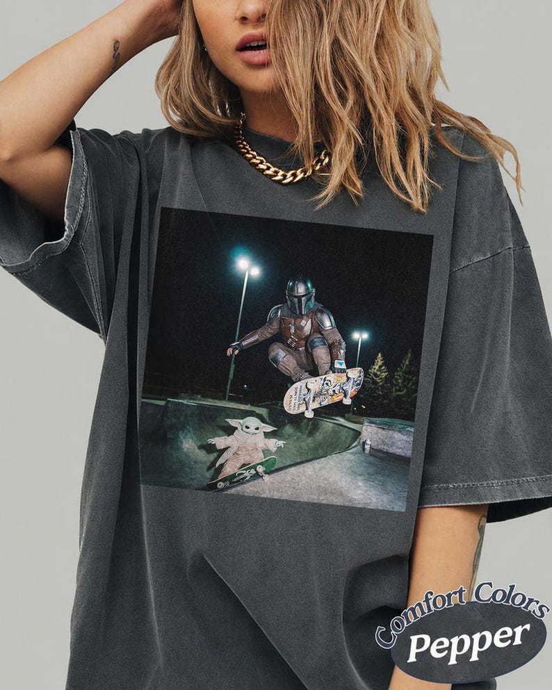 May include: A charcoal gray t-shirt featuring a graphic print of a character in metallic armor skateboarding in the air above a small green creature on a skateboard in a concrete park at night, with the text Comfort Colors Pepper in the corner.