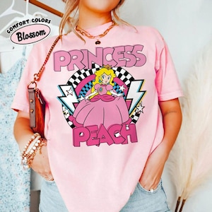 May include: A light pink short-sleeve t-shirt featuring a graphic of Princess Peach in a pink dress. The design includes a black and white checkered circle, pink lightning bolts, and stars. The text reads PRINCESS PEACH in bold pink letters.