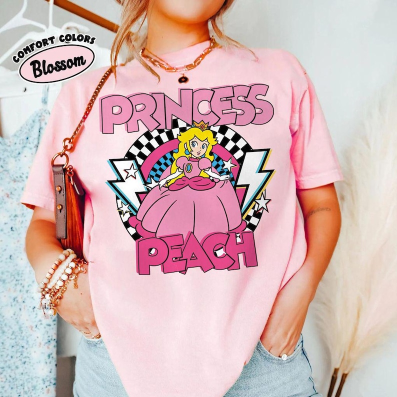 May include: A light pink short-sleeve t-shirt featuring a graphic of Princess Peach in a pink dress. The design includes a black and white checkered circle, pink lightning bolts, and stars. The text reads PRINCESS PEACH in bold pink letters.