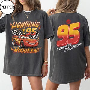 K&ouml;nnte beinhalten: Dunkelgraues T-Shirt mit Lightning McQueen aus dem Film Cars. Auf der Vorderseite sind McQueen, die Nummer 95 und der Text "Lightning McQueen" abgebildet. Auf der R&uuml;ckseite ist eine gro&szlig;e 95 und der Text "Lightning McQueen" zu sehen.
