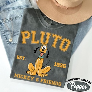 May include: A charcoal gray t-shirt featuring the word "PLUTO" in yellow block letters above a cartoon image of Pluto. The shirt also includes the text "EST. 1926" and "MICKEY & FRIENDS". A tan hat and blue jeans are in the background.