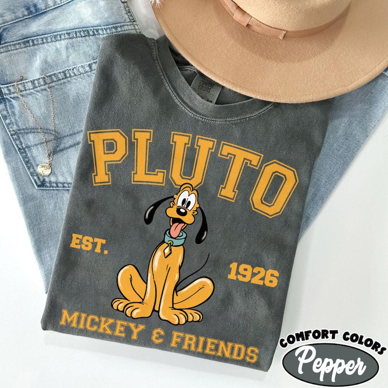 May include: A charcoal gray t-shirt featuring the word "PLUTO" in yellow block letters above a cartoon image of Pluto. The shirt also includes the text "EST. 1926" and "MICKEY & FRIENDS". A tan hat and blue jeans are in the background.