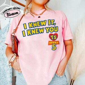 May include: A light pink short-sleeve t-shirt featuring the yellow and blue text I KNEW IT, I KNEW YOU, a red heart with the letters TS, and the word STORY above the number 5 in blue. The shirt has a crew neckline and a relaxed fit.