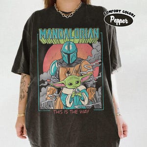 May include: Dark gray t-shirt featuring a graphic of The Mandalorian and Grogu, with the text "MANDALORIAN" and "THIS IS THE WAY". The design includes a red background and a blue and orange color scheme. The shirt has a vintage look.
