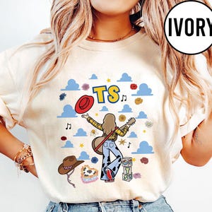 May include: An ivory t-shirt featuring a graphic of a person with long hair playing a guitar, wearing a yellow plaid shirt and cow-print pants. The design includes blue clouds, a red hat, a brown cowboy hat, flowers, music notes, and the text TS.