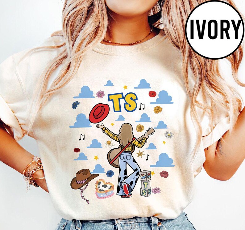 May include: An ivory t-shirt featuring a graphic of a person with long hair playing a guitar, wearing a yellow plaid shirt and cow-print pants. The design includes blue clouds, a red hat, a brown cowboy hat, flowers, music notes, and the text TS.