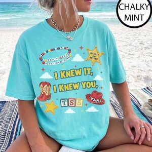 May include: A chalky mint short-sleeve t-shirt featuring a graphic print with the text "I KNEW IT, I KNEW YOU," a beaded bracelet with "You've got a friend in me," a sheriff badge with "INFINITY AND BEYOND," cowboy boots, a hat, and "TS5" blocks.