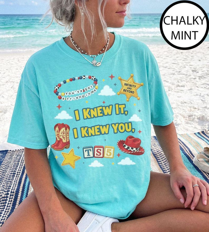 May include: A chalky mint short-sleeve t-shirt featuring a graphic print with the text "I KNEW IT, I KNEW YOU," a beaded bracelet with "You've got a friend in me," a sheriff badge with "INFINITY AND BEYOND," cowboy boots, a hat, and "TS5" blocks.