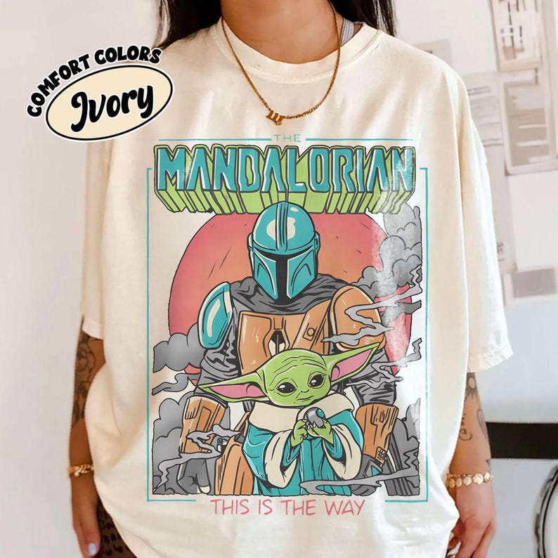 May include: Ivory-colored t-shirt featuring a graphic of The Mandalorian and Grogu, with the text "THE MANDALORIAN" and "THIS IS THE WAY". The design includes a red sun, gray smoke, and a blue and green color scheme.