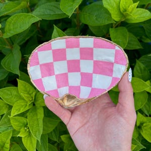 May include: A hand-painted seashell trinket dish. The dish features a pink and white checkered pattern on the inside, with a gold-painted rim and base. The dish is held against a backdrop of green leaves.