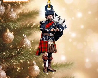 May include: A Christmas tree ornament showing a Scottish bagpiper. The figure wears a kilt in a red and green tartan pattern, a dark blue jacket, and a fur hat. The text "NAME CAN BE CHANGED" is at the top. The ornament is suspended by a gold string.