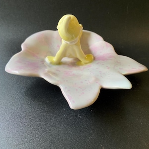 May include: A ceramic trinket dish shaped like a leaf with a pale pink speckled finish. A small, yellow, gnome-like figure with a pointed hat is attached to the center of the dish, sitting with legs extended forward on the surface of the leaf.