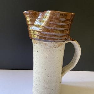 May include: A tall ceramic pitcher featuring a speckled off-white cylindrical body and a contrasting dark brown glazed rim. The vessel includes a curved handle on the side and a wide, flared spout at the top, showcasing a distinct two-tone finish.