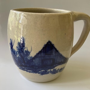 May include: A beige ceramic mug with a speckled texture and a handle on the right side. The exterior features a dark blue transferware-style illustration of a barn surrounded by trees, wrapping around the lower portion of the rounded vessel.