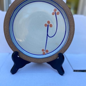 May include: A handmade ceramic plate with a cream-colored center, adorned with a blue and orange floral design. The plate has a brown rim and is displayed on a black stand. The plate's diameter is approximately 10 inches.