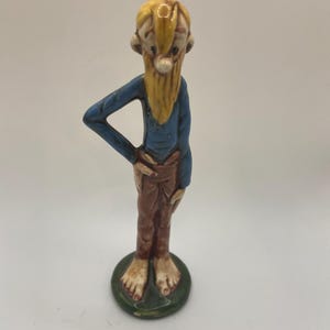 May include: A ceramic figurine of a bearded man with a long yellow beard and hair, wearing a blue shirt and brown pants. The figurine is standing on a green circular base. The man has his hand on his hip.