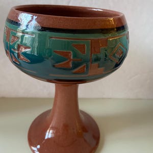 May include: A brown ceramic goblet with a green and brown geometric design. The goblet has a wide bowl and a tall, slender stem.