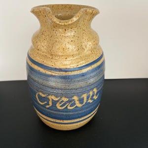 May include: A handmade ceramic creamer with a tan speckled finish and blue stripes. The word "cream" is written in cursive on the blue band. The creamer has a flared top and a spout for easy pouring. A rustic and charming addition to any kitchen.