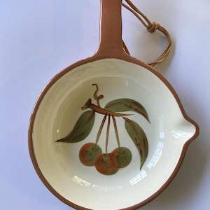 May include: Ceramic serving dish with a brown handle and rim, featuring a painted cherry design. The dish has a cream-colored interior and a small spout. A leather cord is attached for hanging.