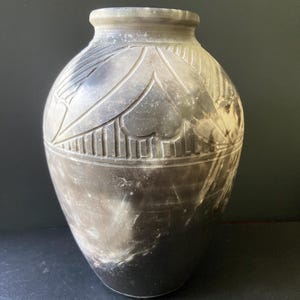 May include: A rounded stone vase featuring a mottled dark gray and white surface with a smooth finish. The upper section is decorated with carved, symmetrical heart-shaped motifs and vertical line patterns encircling the neck of the vessel.