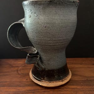 May include: A handmade ceramic mug with a unique, sculpted handle. The mug features a dark gray glaze with a textured surface and a contrasting dark brown base. The mug is approximately 4 inches tall.