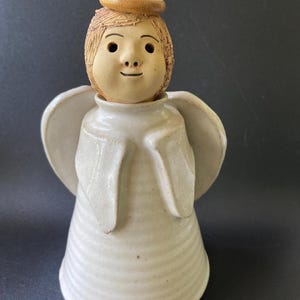 May include: A ceramic angel figurine featuring a light beige unglazed head with a matching halo, set atop a white glazed, ribbed conical body. The figure has carved facial features, including eyes, a nose, and a small mouth, with two wings.