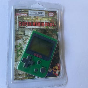 May include: A green Mini Classics Super Mario Bros. handheld game console in a clear plastic package. The console has a black directional pad and buttons. The packaging includes the Nintendo logo and the text "Where are you Princess Toadstool?"
