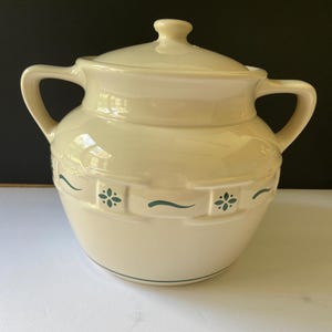 May include: A cream-colored ceramic sugar bowl with a matching lid and two side handles. The exterior features a raised basket-weave texture around the center, accented with small green floral motifs and thin horizontal green lines.