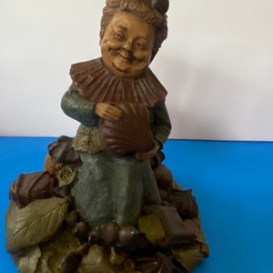 May include: A ceramic figurine of a woman with a whimsical expression, wearing a brown and green dress and a brown hat. She is surrounded by chocolate-colored objects, including leaves and acorns.