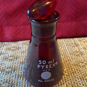 May include: A red Pyrex laboratory flask with a matching stopper. The flask is marked with "50 ml PYREX USA" and the number "55020". The flask is made of glass and has a conical shape.