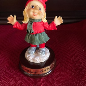 May include: A figurine of a smiling, blonde-haired girl wearing a red hat, jacket, and boots, with a green scarf and skirt. The figurine is standing on a snowy base atop a dark brown base with gold lettering. The figurine has its arms outstretched.