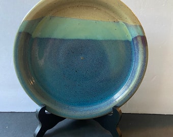 May include: A ceramic plate with a blue, green, and beige glaze. The plate is on a black stand.