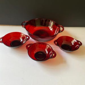 May include: A set of four ruby red glass bowls featuring a large central bowl and three smaller matching bowls. Each bowl has two side handles and a vertical ribbed pattern along the exterior base, crafted from translucent red glass.