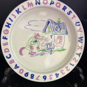 May include: White ceramic plate with a colorful illustration of a wolf and a house. The plate features the alphabet in purple and blue around the rim, with numbers at the bottom. The wolf is wearing a green suit and the house has a blue roof.