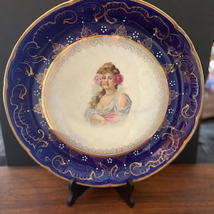 May include: A decorative porcelain plate with a dark blue border and gold accents. The center features a portrait of a woman with flowers in her hair. The plate is displayed on a black stand, showcasing its elegant design.