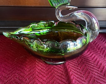 May include: A decorative glass swan-shaped bowl. The bowl is a deep green color with a clear, curved neck and a green leaf-shaped accent. The bowl sits on a red patterned surface.