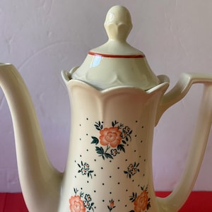 May include: A white ceramic coffee pot with a floral pattern. The pot has a lid with a knob on top. The floral pattern features orange roses and green leaves on a white background with small white dots.